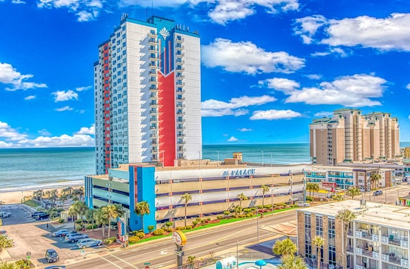 A high-rise hotel with a prominent red and blue facade, a parking lot, and a view of the ocean.