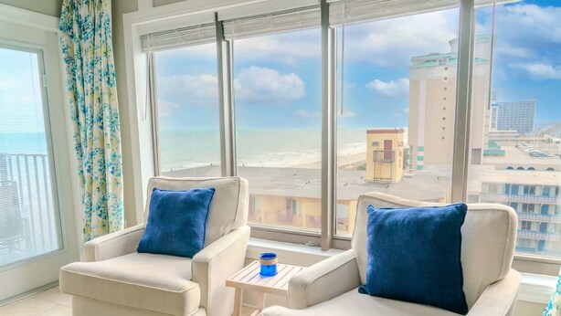 Ein Zimmer mit zwei Sesseln, einem kleinen Tisch und Blick auf den Strand und die Gebäude.