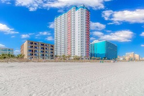 A beachfront hotel with multiple floors, balconies, and a sandy beach in the foreground.