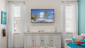 Een woonkamer met een witte kast, een televisie waarop een strand te zien is, en een schaal glazen decoraties op een tafel.