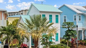 Een rij kleurrijke huizen, met in het midden een wit huis met een groen dak en turquoise luiken, omgeven door weelderige beplanting en palmbomen.