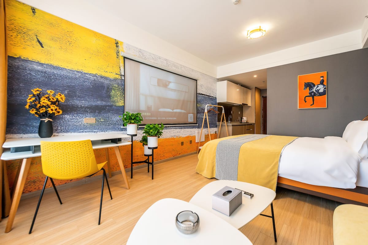 A modern hotel room with a large bed, a desk with a chair, a small table, and a potted plant.