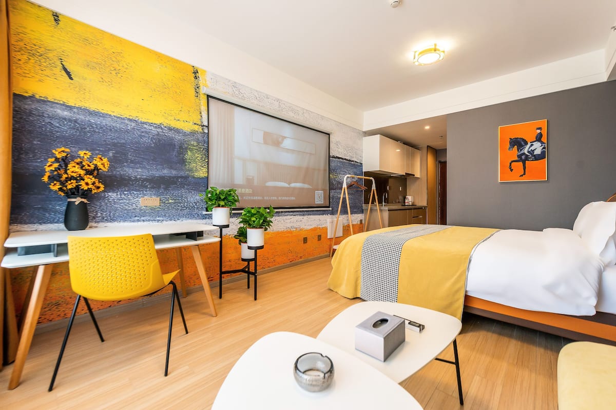 A modern hotel room with a bed, a desk, a chair, a small table, and a large abstract painting on the wall.