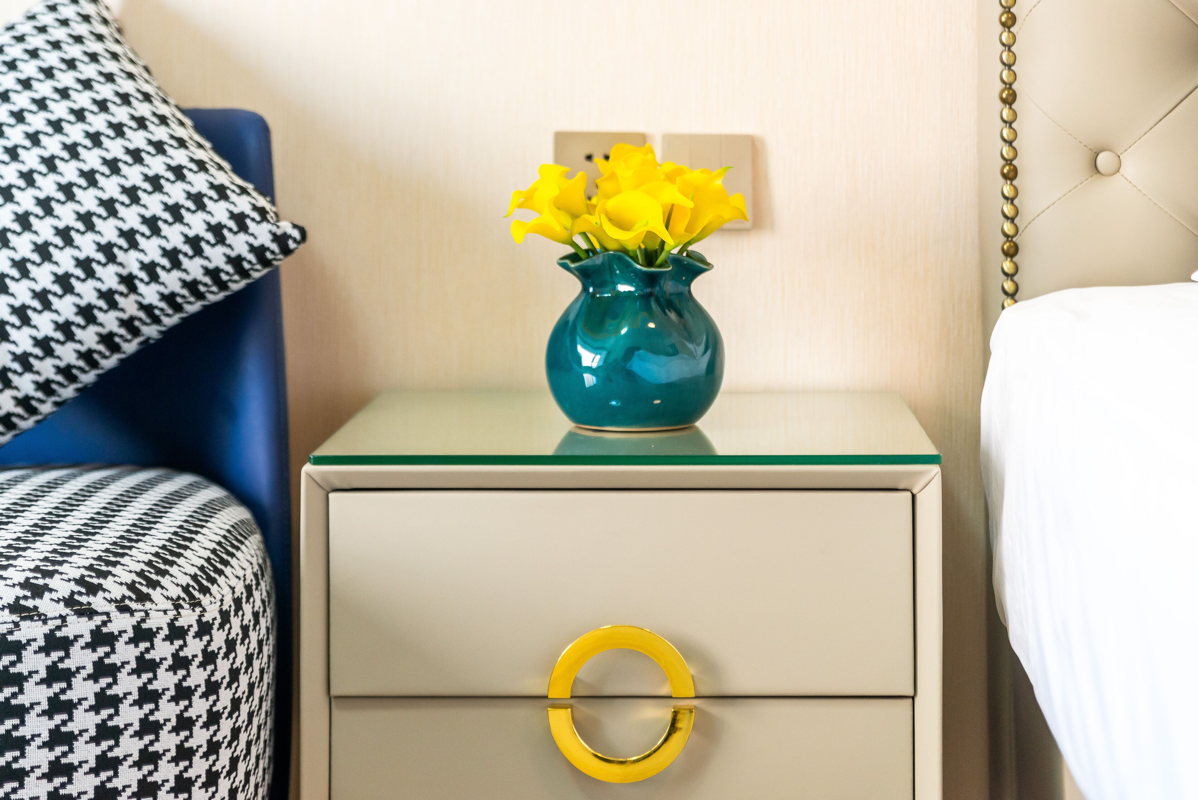A nightstand with a teal vase of yellow flowers, a houndstooth pillow, and a bed with white bedding.