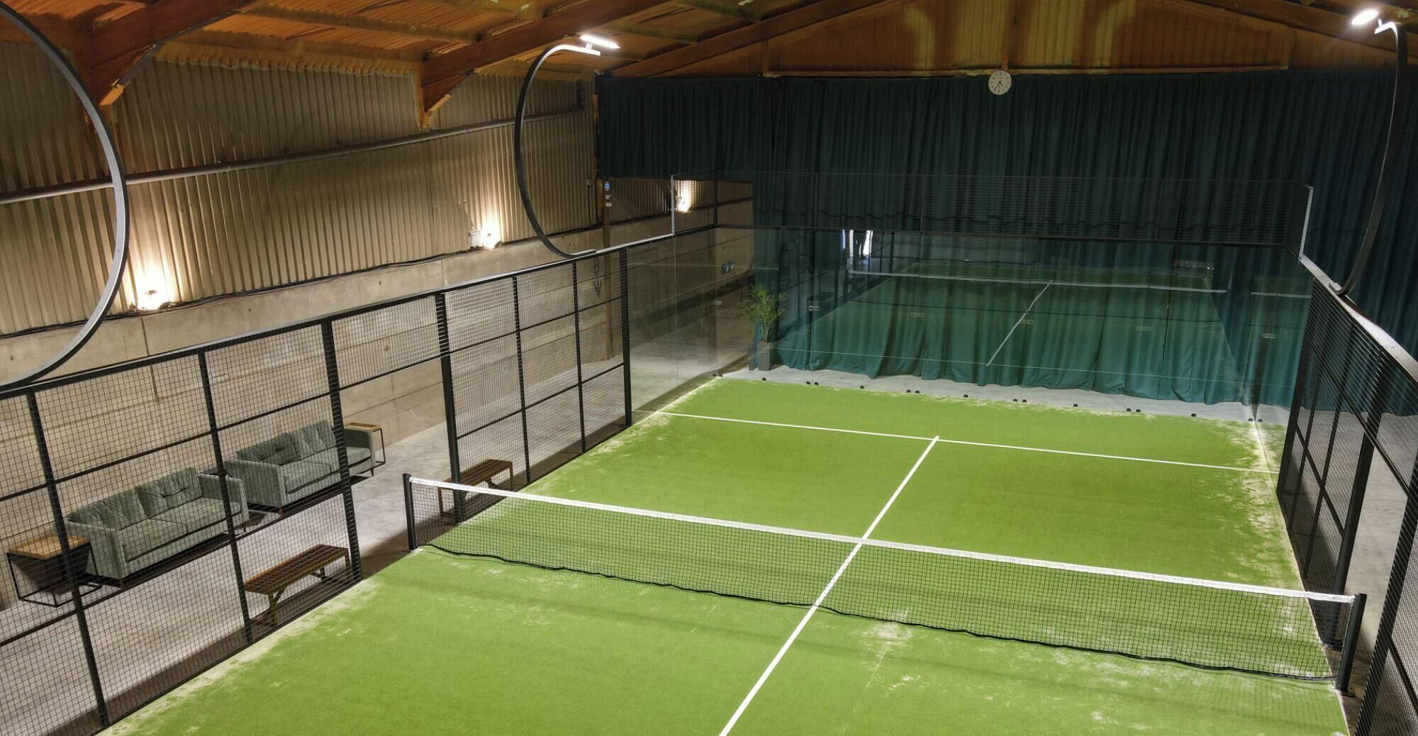 Un court de tennis intérieur doté d’un filet, d’une zone pour les spectateurs et d’un espace clos avec un plafond en bois.