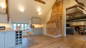 A spacious loft with exposed wooden beams, a brick fireplace, and a kitchen with white cabinets and a bar area. - Apple Loft -  Beautiful Somerset cottage, sleeps 13 guests  in 6 bedrooms (Ilminster)