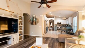 A spacious living room with a flat-screen TV, wooden entertainment unit, and a ceiling fan. A dining area with a kitchen in the background is visible.