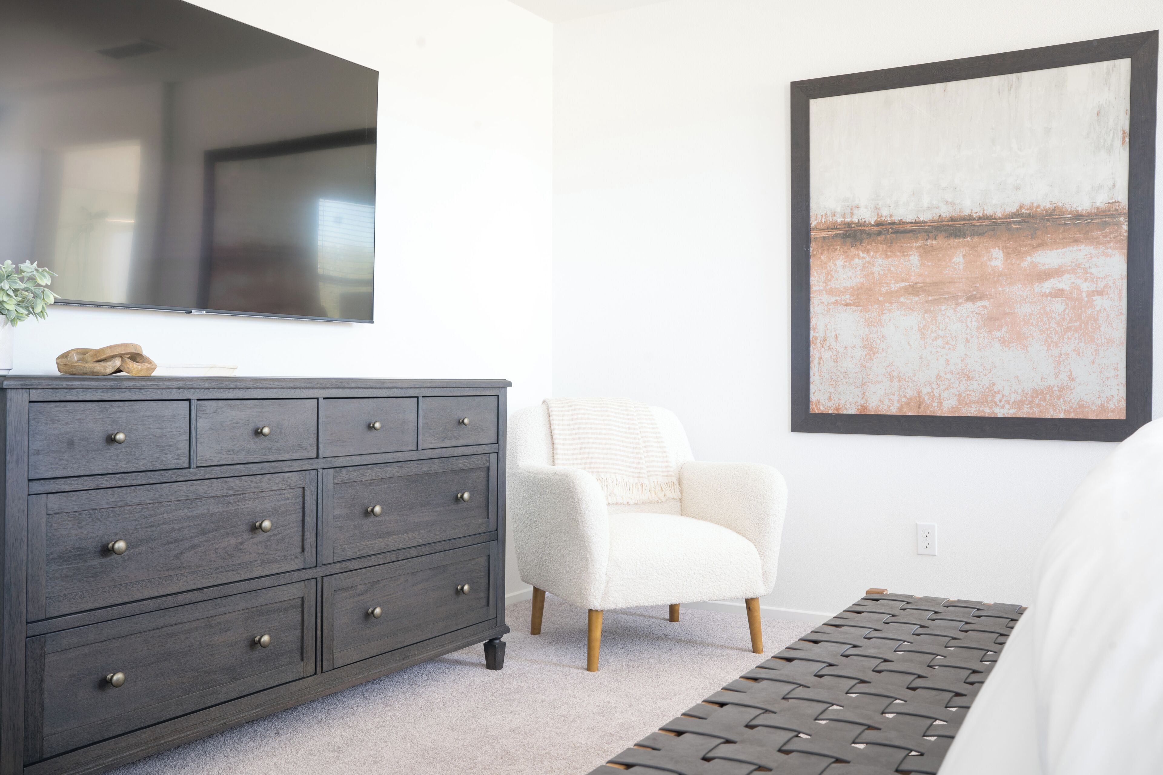 Slaapkamer met een donker houten dressoir, een witte fauteuil, een groot abstract schilderij en een kamerkleed met dessin.