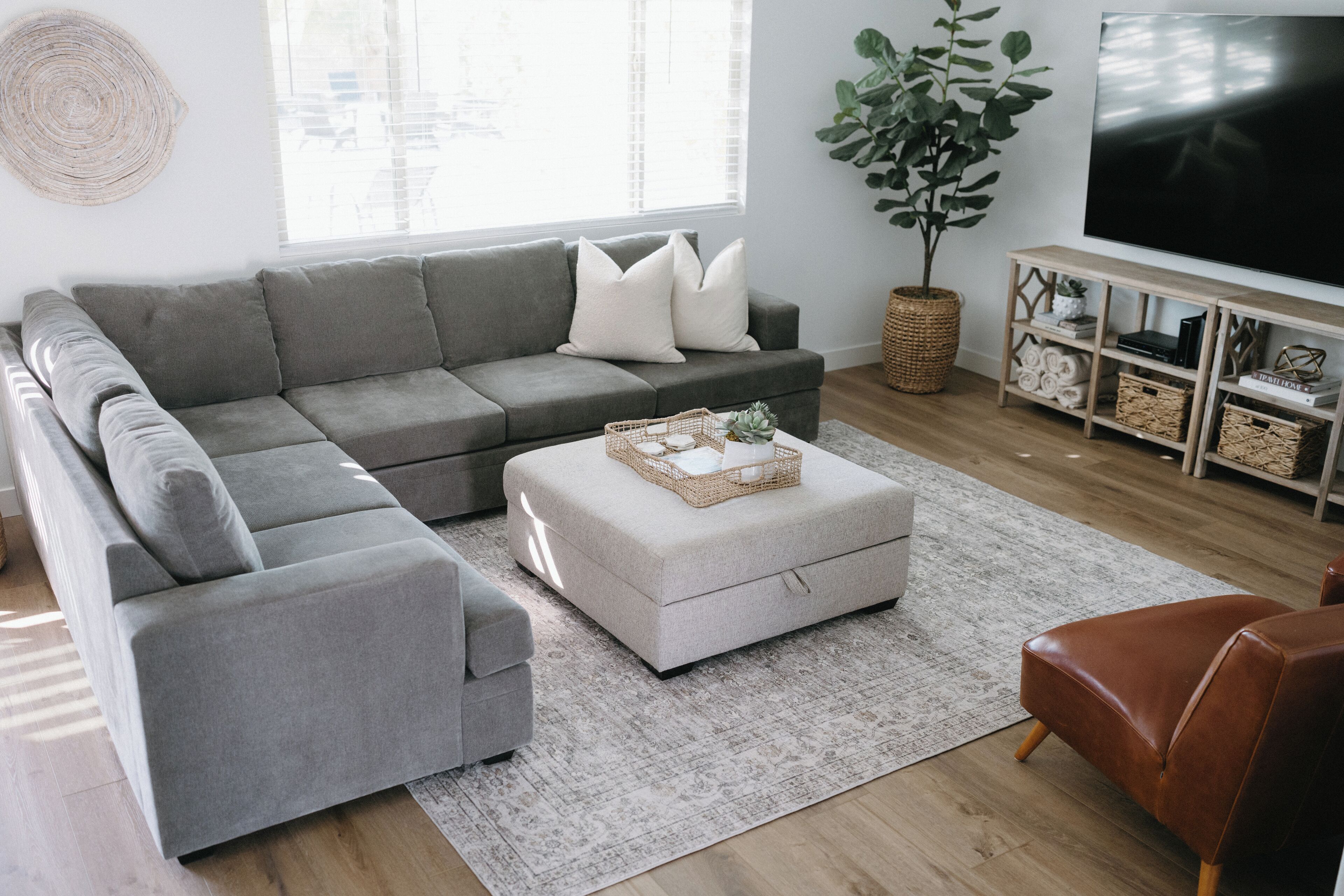 Un salon moderne avec un canapé d’angle gris, un pouf blanc, un fauteuil en cuir marron, une grande télévision et une plante en pot.
