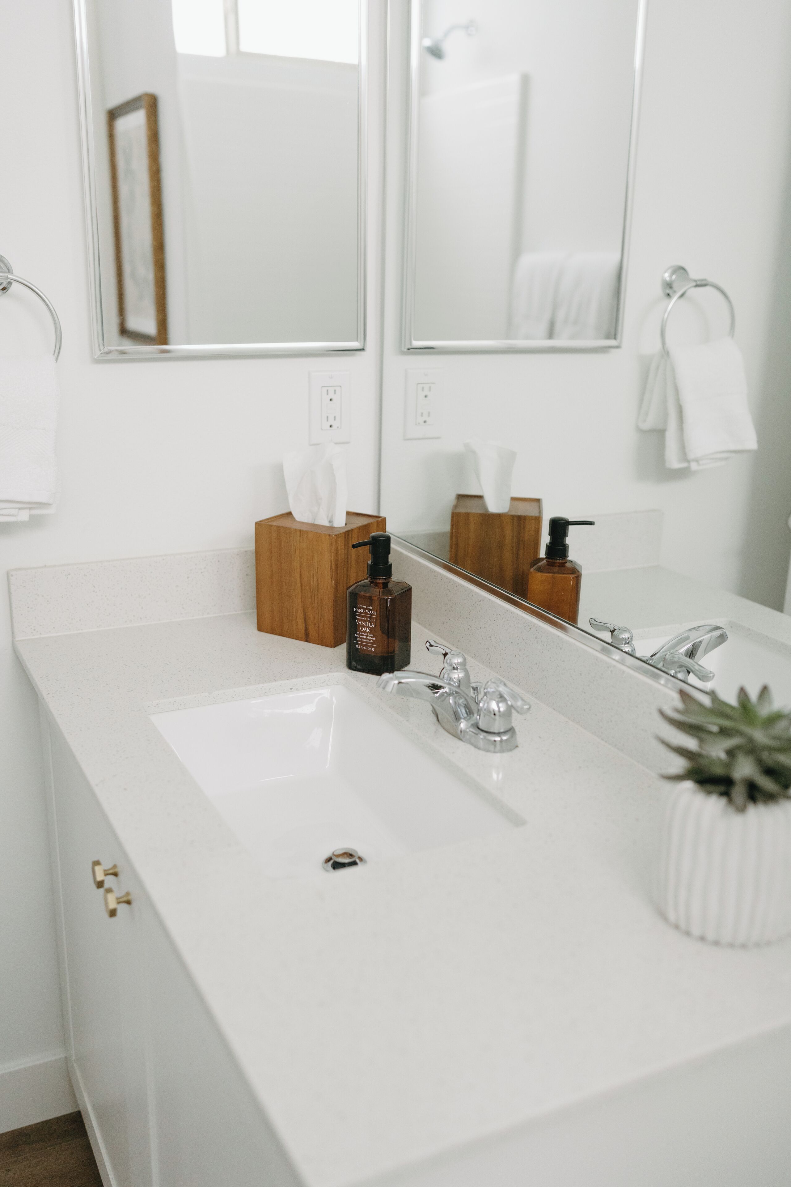 A modern bathroom with a white sink, two mirrors, a wooden soap dispenser, and a potted plant.