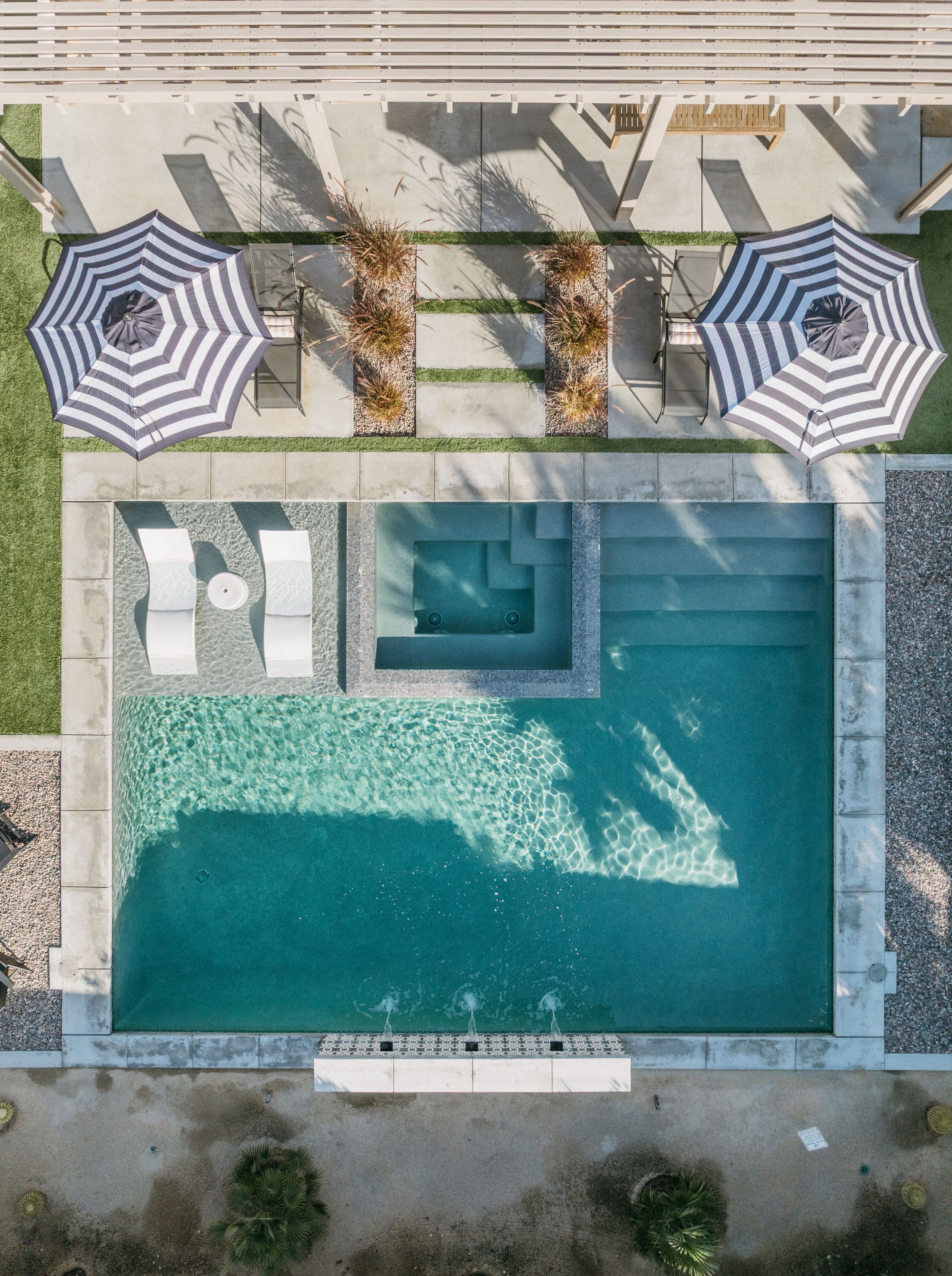 Vista aérea de una piscina con dos sillas reclinables y dos sombrillas a rayas, rodeada por una terraza y un pequeño jardín.