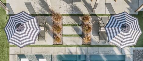 Vista aérea de una piscina con dos sillas reclinables y dos sombrillas a rayas, rodeada por una terraza y un pequeño jardín.