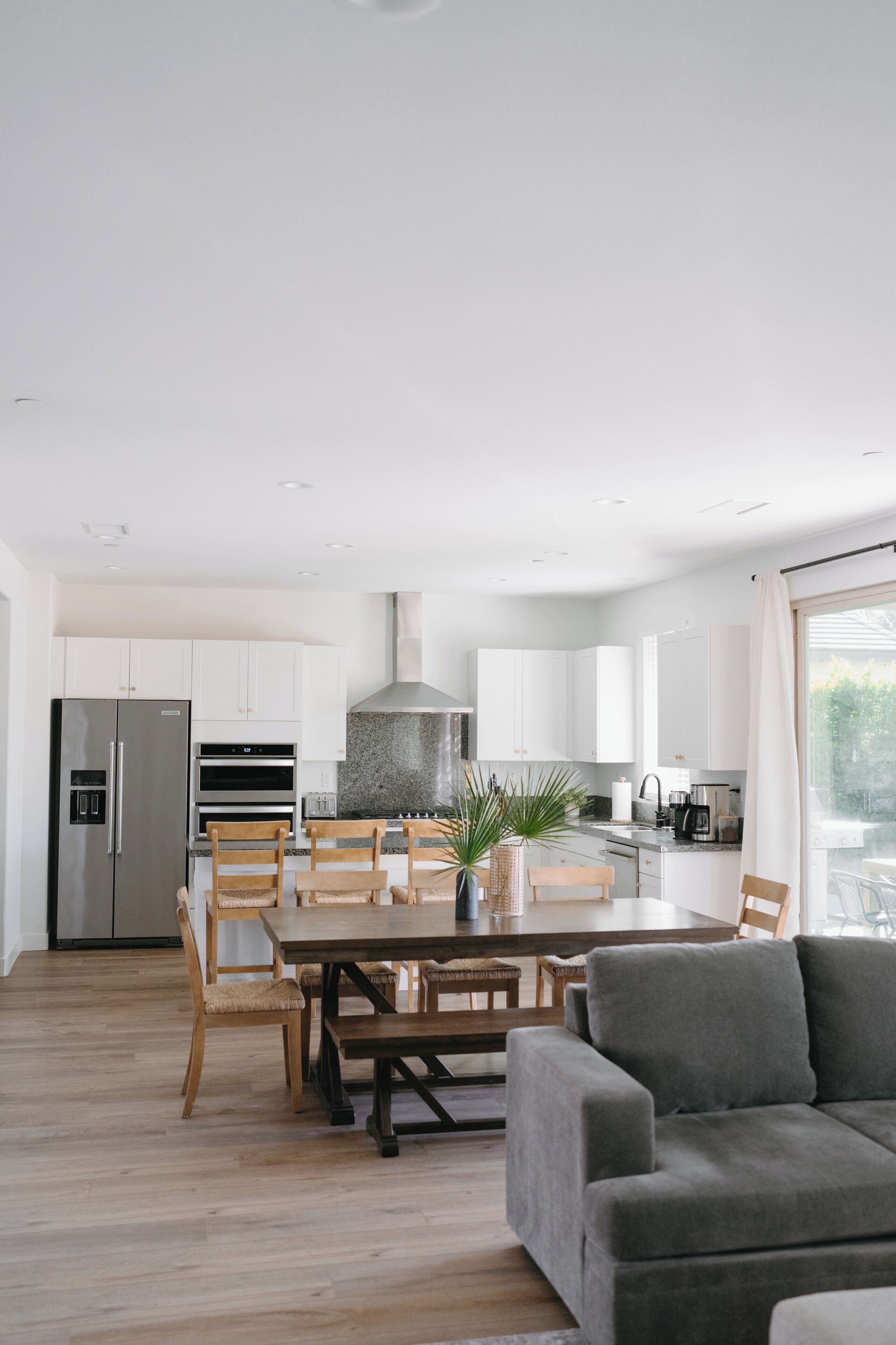 A modern kitchen with white cabinets, a stainless steel refrigerator, and a dining area with wooden chairs and a grey sofa.