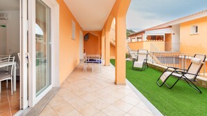 Un balcone con una porta vetrata, un tavolo con sedie e una zona verde con poltroncine.