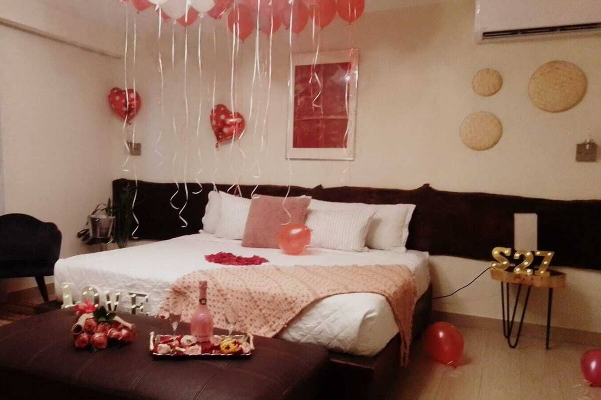 Ein Hotelzimmer mit einem Bett, einem kleinen Tisch und einem Stuhl. Auf dem Bett liegen Ballons und ein Blumenstrauß.