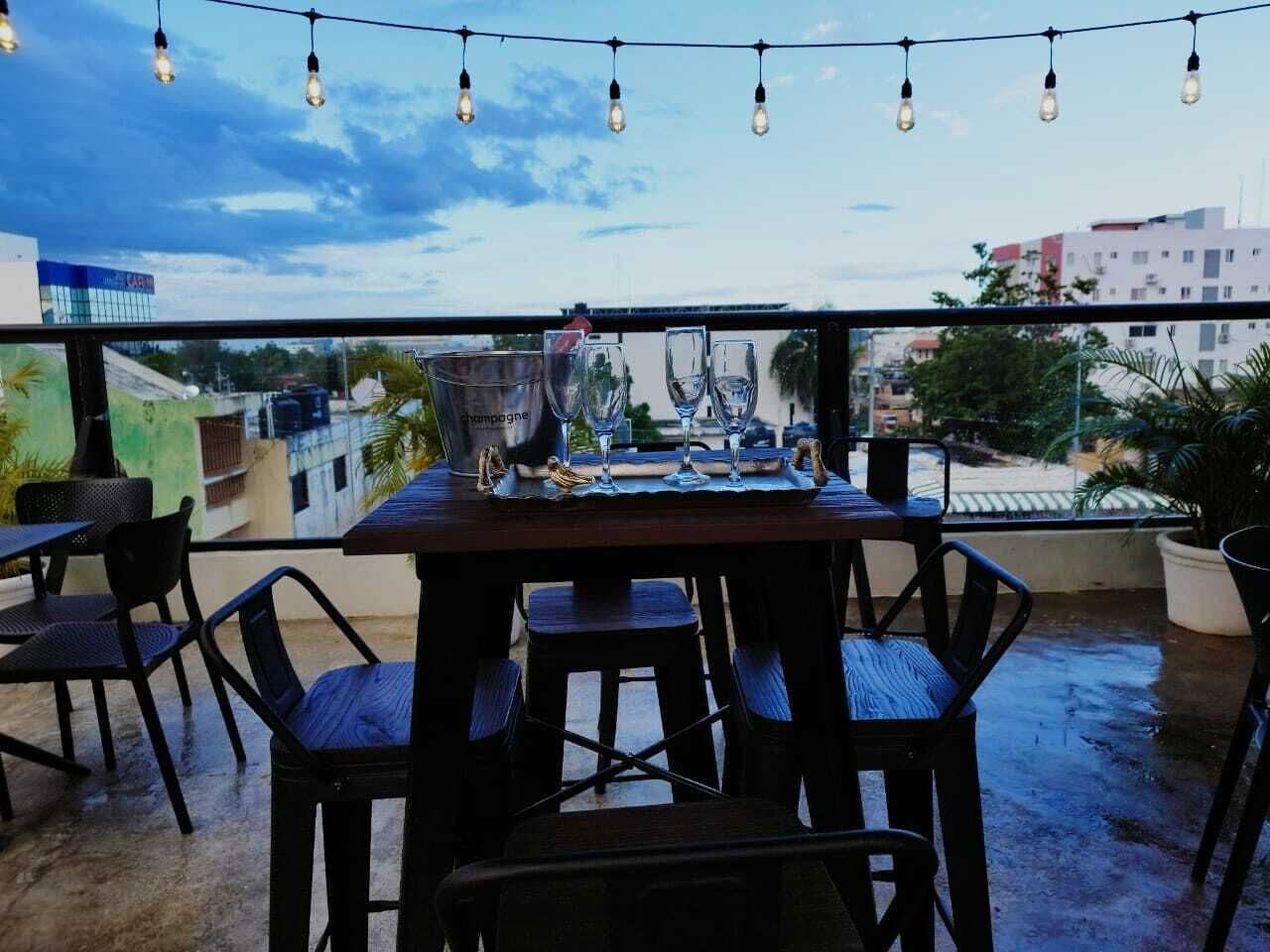An outdoor dining area with a table and chairs, string lights overhead, and a view of buildings in the background.