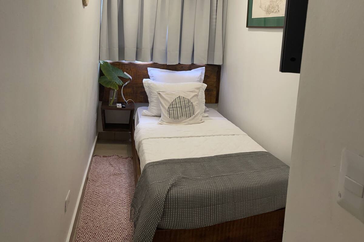 Ein kleines, ordentlich eingerichtetes Schlafzimmer mit einem Einzelbett, einem Nachttisch, einer Zimmerpflanze und einem gerahmten Bild an der Wand.