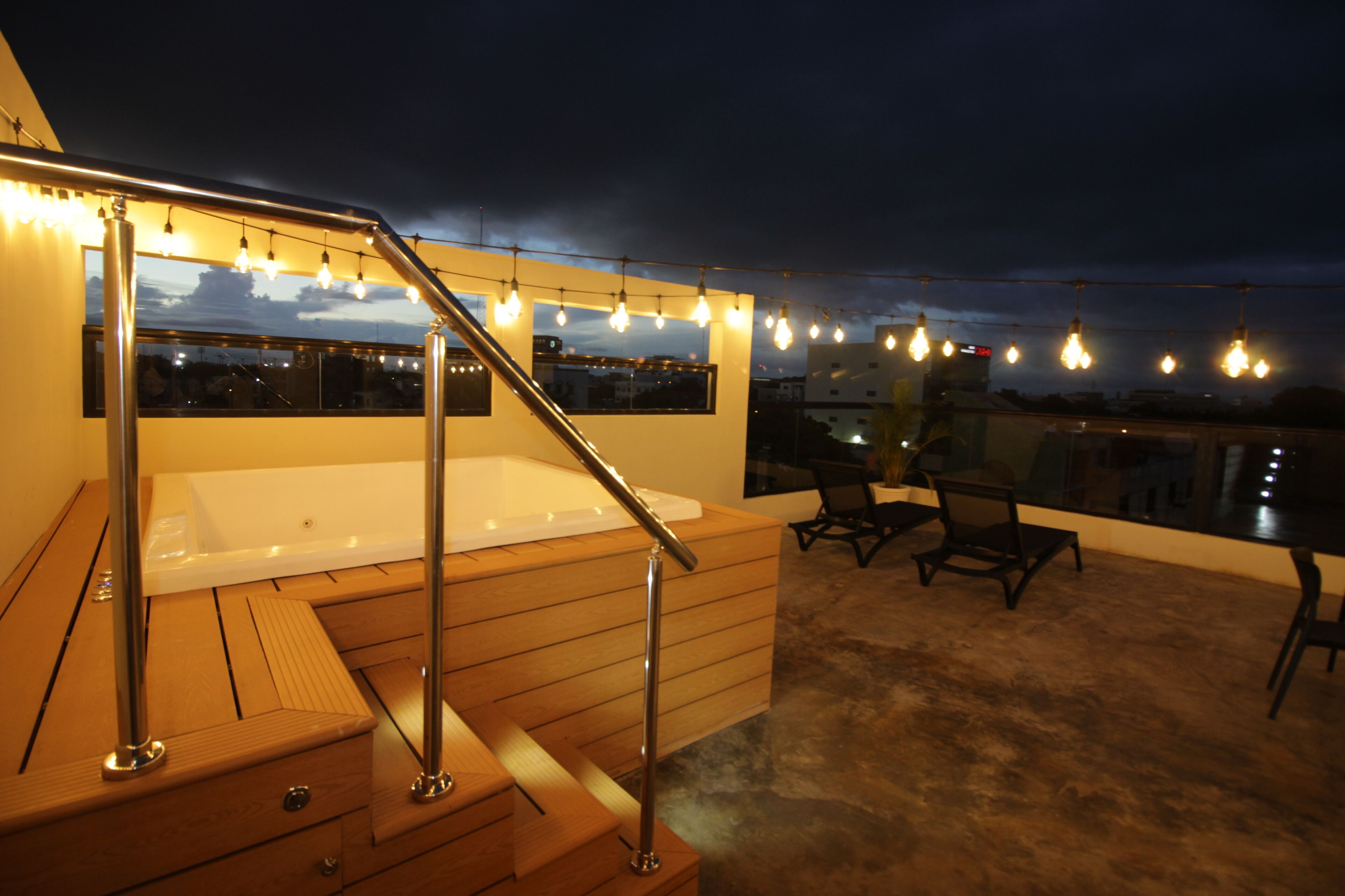 A rooftop terrace with a hot tub, wooden flooring, metal railings, and string lights.