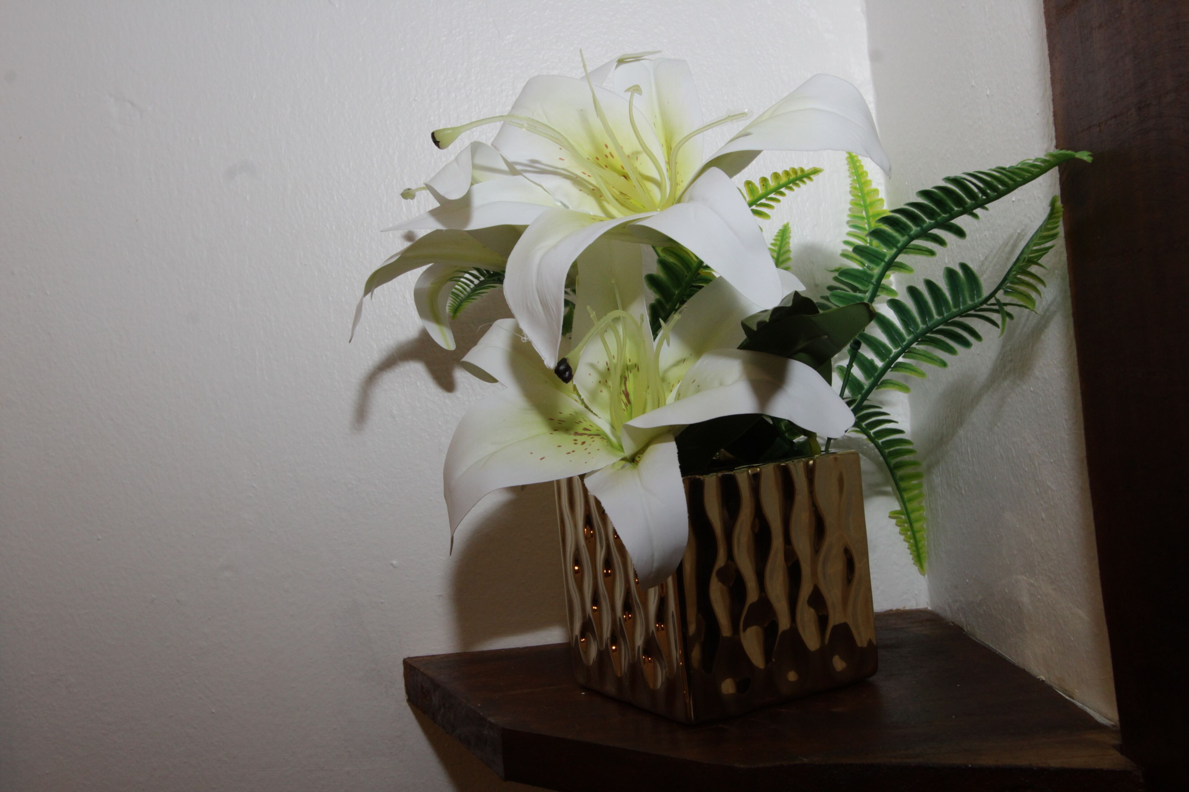 Eine weiße Lilie in einer dekorativen Vase auf einem Holzregal vor einer weißen Wand.