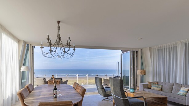 A spacious living area with a large round table, comfortable seating, and a view of the beach.