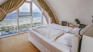 A bedroom with a large window overlooking a beach, a bed, a chair, and a desk with a lamp.