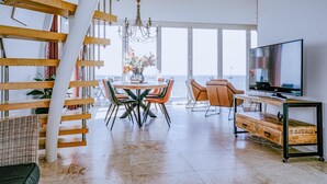 A spacious living area with a high ceiling, a wooden staircase, a dining table with chairs, a television, and a view of the sea.