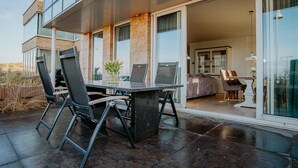 Un patio al aire libre con una mesa y sillas, un ventanal de vidrio que da a una zona de estar interior y una fachada de edificio moderna.
