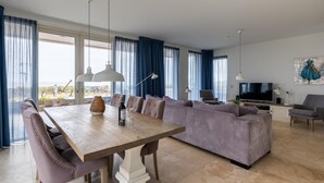 Una sala de estar moderna con una mesa de comedor de madera, asientos acolchados y ventanas grandes con cortinas azules.