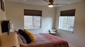 Un dormitorio con cama, dos ventanas, ventilador de techo y mesita de noche.