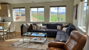 Una sala de estar moderna con un sofá modular, una mesa de centro de vidrio y un sillón de cuero. Ventanas amplias ofrecen vista a un campo verde y montañas distantes.