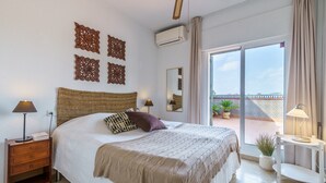 Ein Schlafzimmer mit Bett, Nachttischen, einem Fenster und einem Balkon mit Pflanzen.