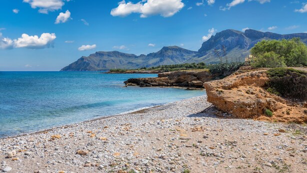 A coastal landscape with a rocky beach, clear blue sea, and mountains in the background.