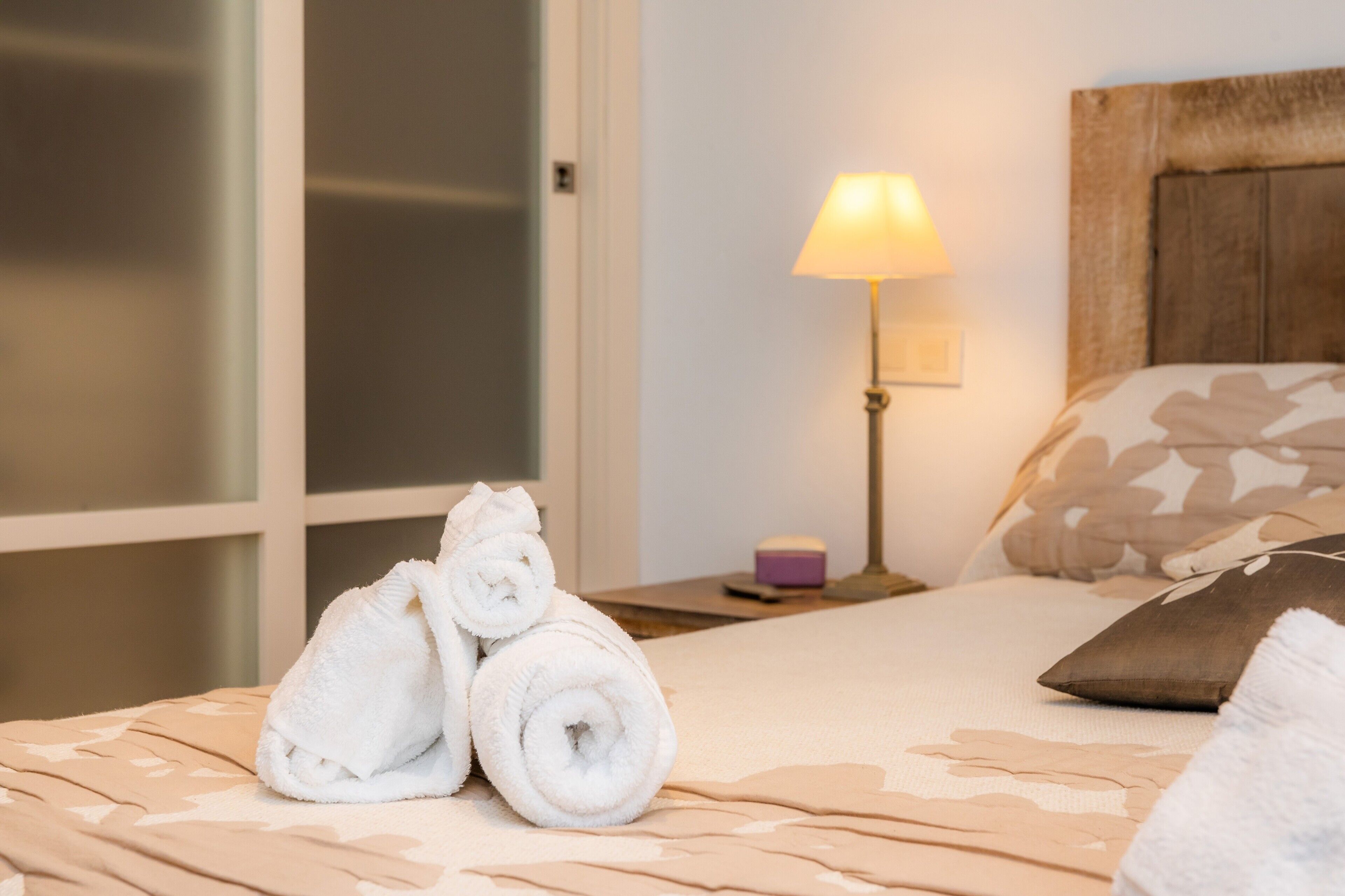 Een net opgemaakt bed met opgerolde handdoeken, een bedlamp en een nachtkastje met een klein object.