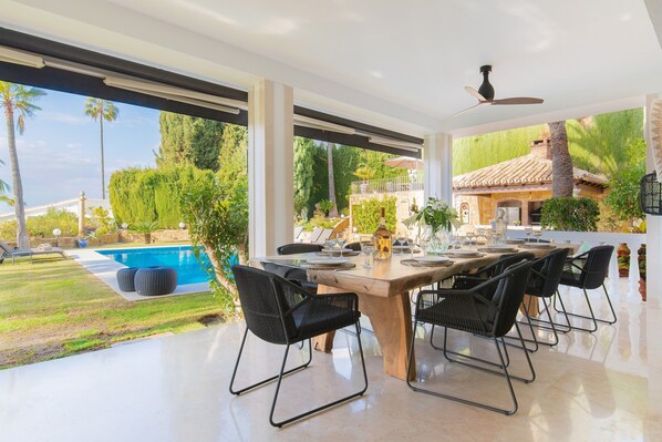 A modern outdoor dining area with a pool, chairs, and a view of a villa.
