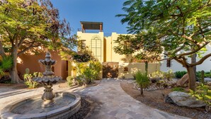 A courtyard with a stone pathway leading to a building, surrounded by various plants and a central fountain. - Casa Juanchi by Casago (Loreto)