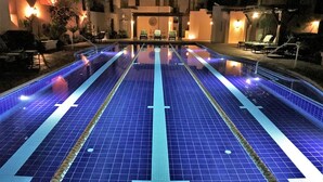 A swimming pool with blue tiles and white lane markings, surrounded by lounge chairs and a building with warm lighting. - Casa Juanchi by Casago (Loreto)