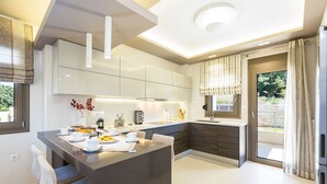 A modern kitchen with a central island, built-in cabinets, and a large window with curtains. - Luxury Villa set in nature with private pool (Rethymno)