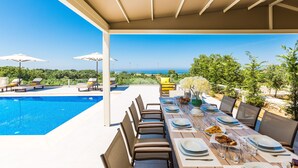 A dining area with a pool view, outdoor seating, and a clear sky. - Luxury Villa set in nature with private pool (Rethymno)
