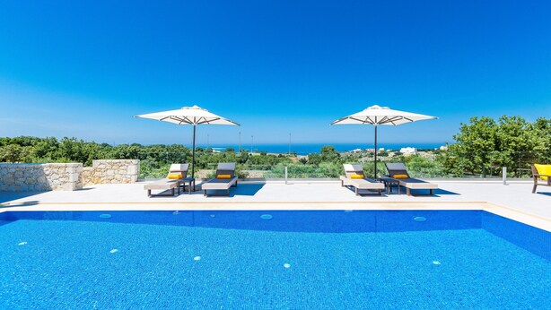 A swimming pool with lounge chairs and umbrellas, surrounded by greenery and a clear view of the sea. - Luxury Villa set in nature with private pool (Rethymno)