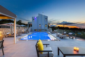 A modern pool area with lounge chairs and umbrellas, a stone wall, and a view of the sunset.