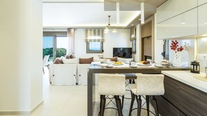A modern kitchen with a central island, bar stools, and a dining area with a sofa and table. - Luxury Villa set in nature with private pool (Rethymno)