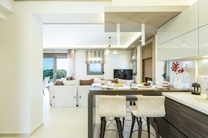 A modern kitchen with a central island, bar stools, and a dining area with a sofa and table. - Luxury Villa set in nature with private pool (Rethymno)