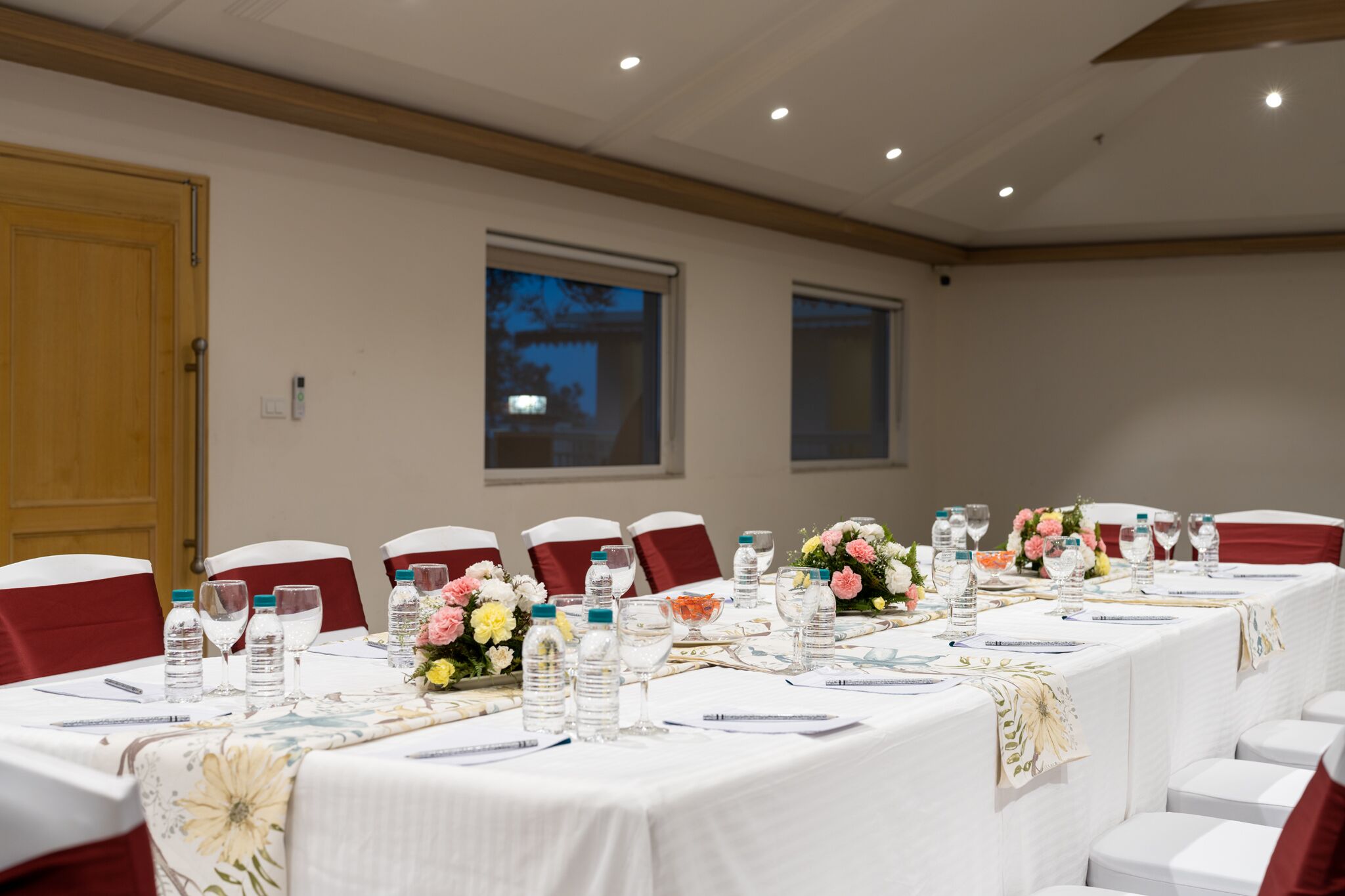 A conference room with a long table set for a meeting, equipped with water bottles, glasses, and floral centerpieces.