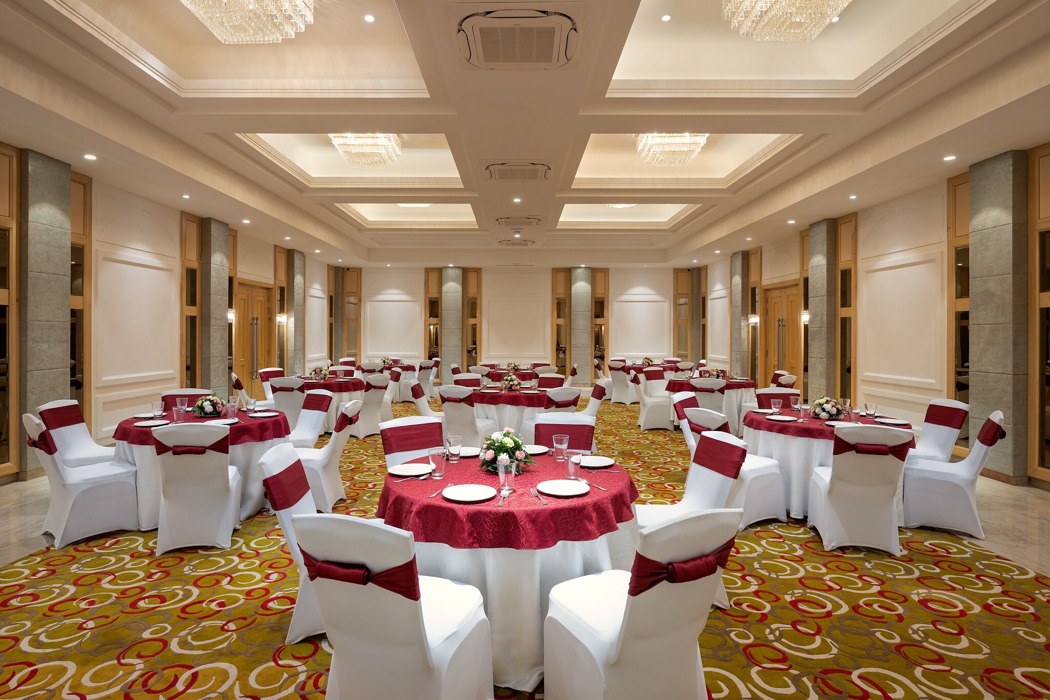 Un salón de banquetes con mesas redondas, sillas blancas y cintas rojas.