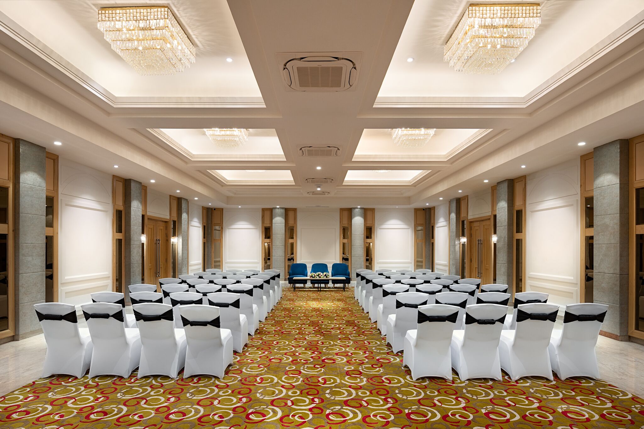 Une salle de conférence avec des rangées de chaises blanches, une moquette à motifs, des lustres et un canapé bleu.