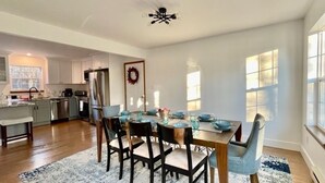Un vaste espace dédié à la salle à manger, avec une table et des chaises en bois, une cuisine à l’arrière-plan et de grandes fenêtres qui laissent entrer la lumière naturelle.