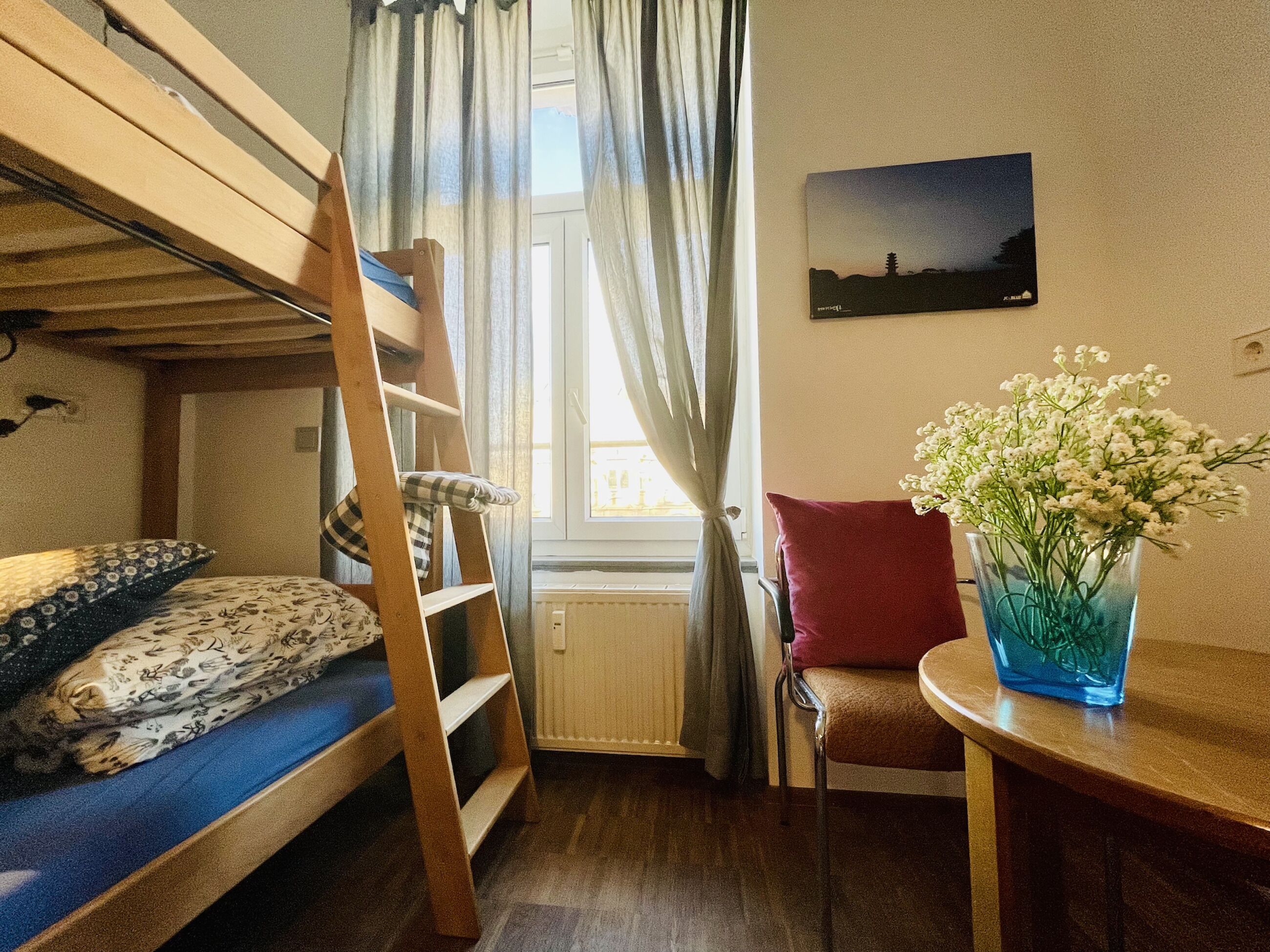 A room with a bunk bed, a chair, a table, a vase with flowers, and a window with curtains.