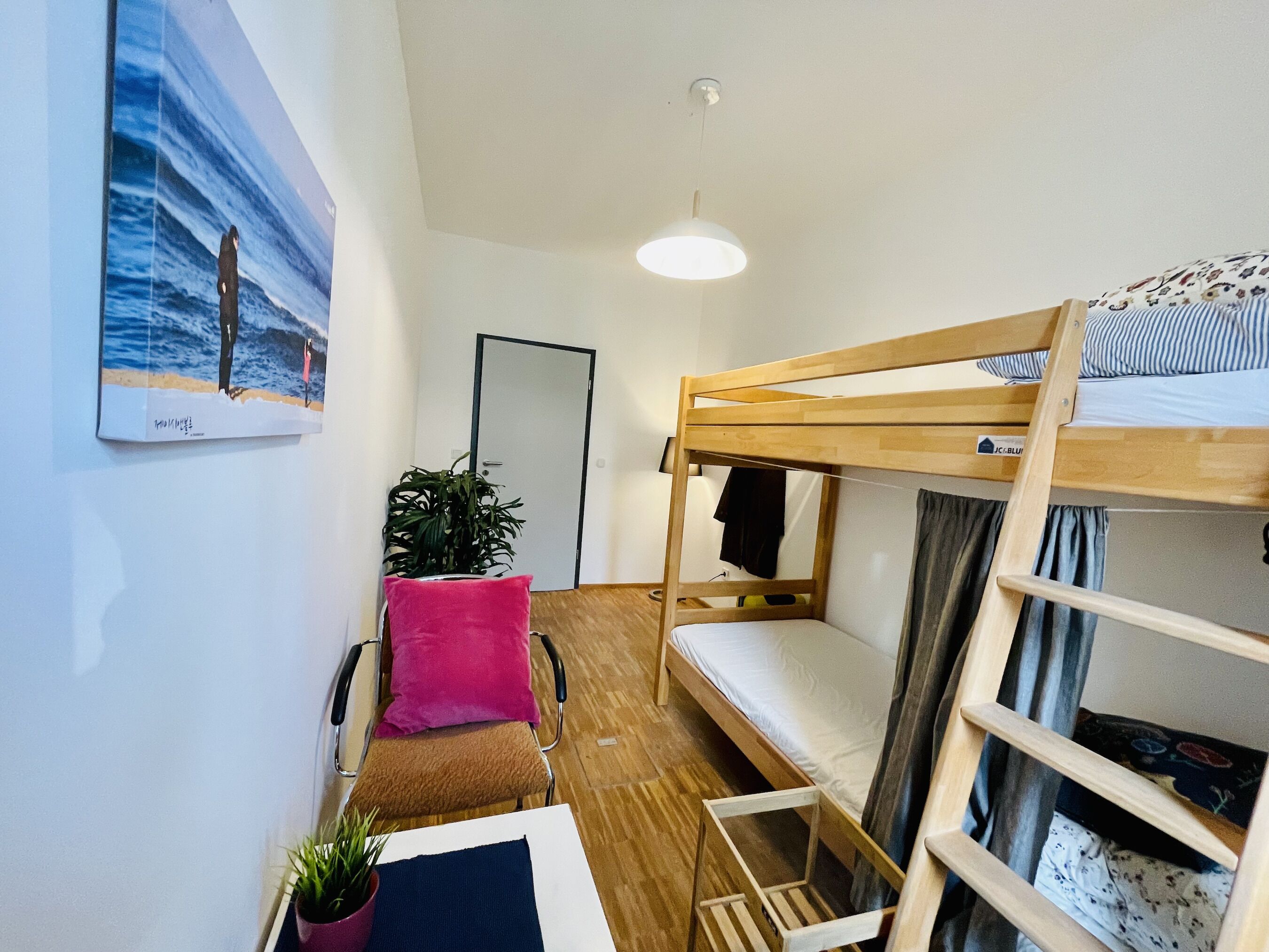 A bunk bed room with a wooden floor, a desk, a chair, a pink pillow, a potted plant, and a painting of a beach scene.