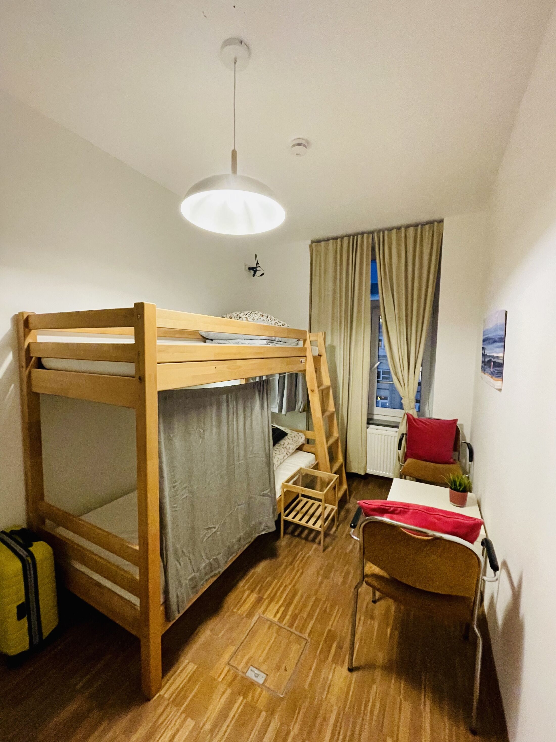 Una cama litera con escalera, una mesita con una planta, una silla y una ventana con cortinas.