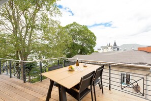 Une terrasse en bois avec une table et des chaises, d’où l’on peut admirer une vue sur la ville, avec ses bâtiments et son église.